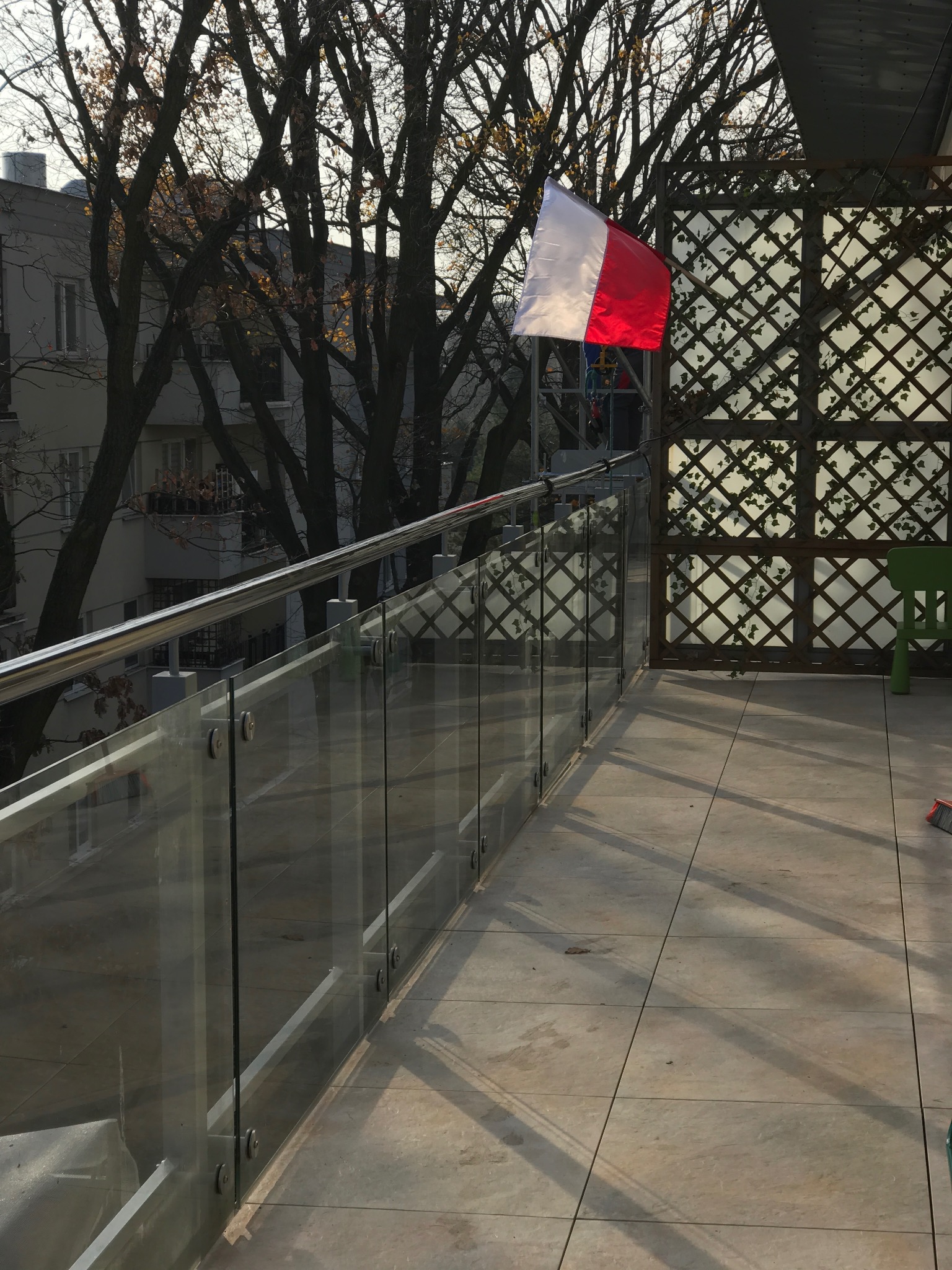 Nowoczesny balkon z szklaną balustradą i płytkami. W tle budynek mieszkalny i drzewa. Na balkonie flaga Polski i zielone krzesło. Widok z góry, oświetlenie dzienne.