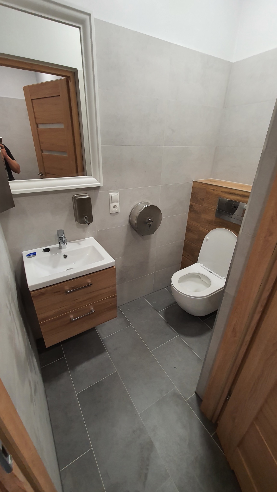 Wykończona łazienka w szarościach z drewnianymi akcentami: umywalka z szafką, toaleta podwieszana i lustro w białej ramie. Widoczny dozownik mydła i podajnik ręczników.