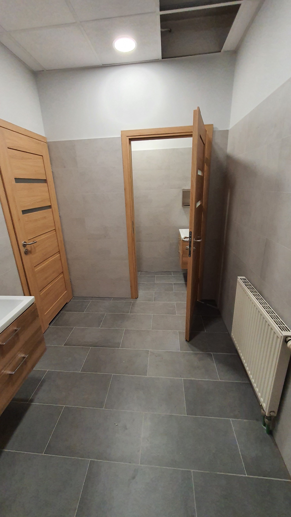 Wykończona łazienka z szarą płytką podłogową, drewnianymi drzwiami i grzejnikiem. Widok na umywalkę w głębi. Częściowo widoczny sufit podwieszany.