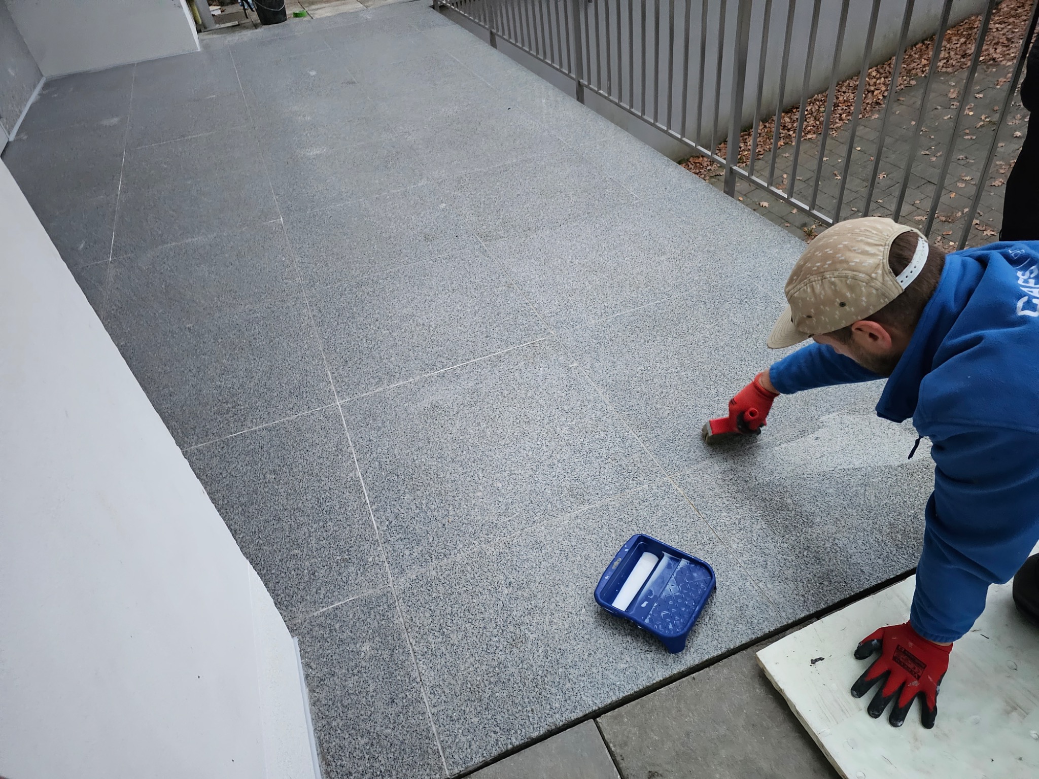 Pracownik w niebieskiej bluzie impregnuje szarą, kwadratową płytkę na balkonie. Widoczny pędzel, kuweta malarska i fragment balustrady.