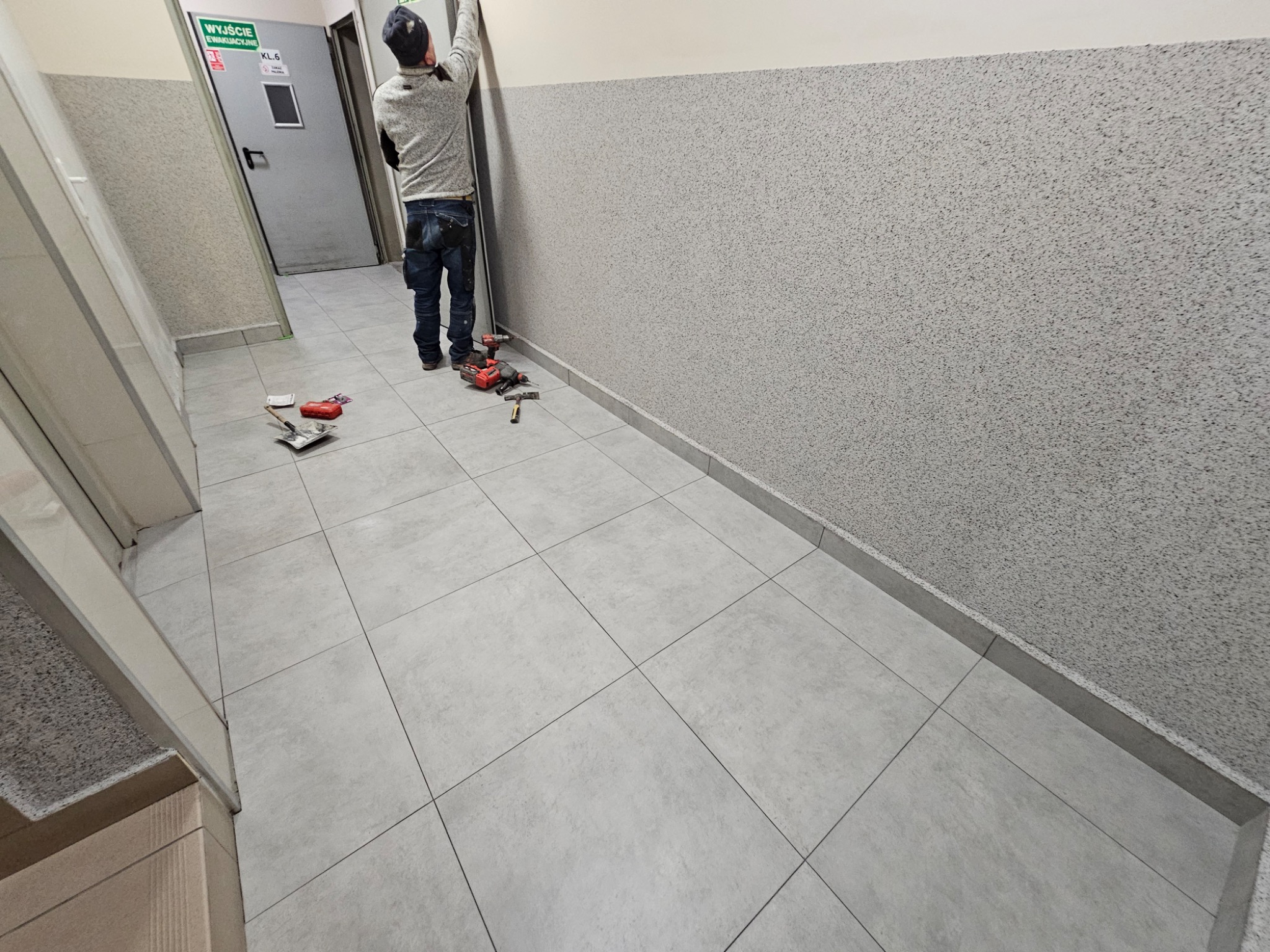 Pracownik w trakcie montażu listwy przypodłogowej w korytarzu. Widoczne narzędzia: wiertarka, młotek. Ściany wykończone szarym tynkiem strukturalnym, podłoga wyłożona szarymi płytkami.