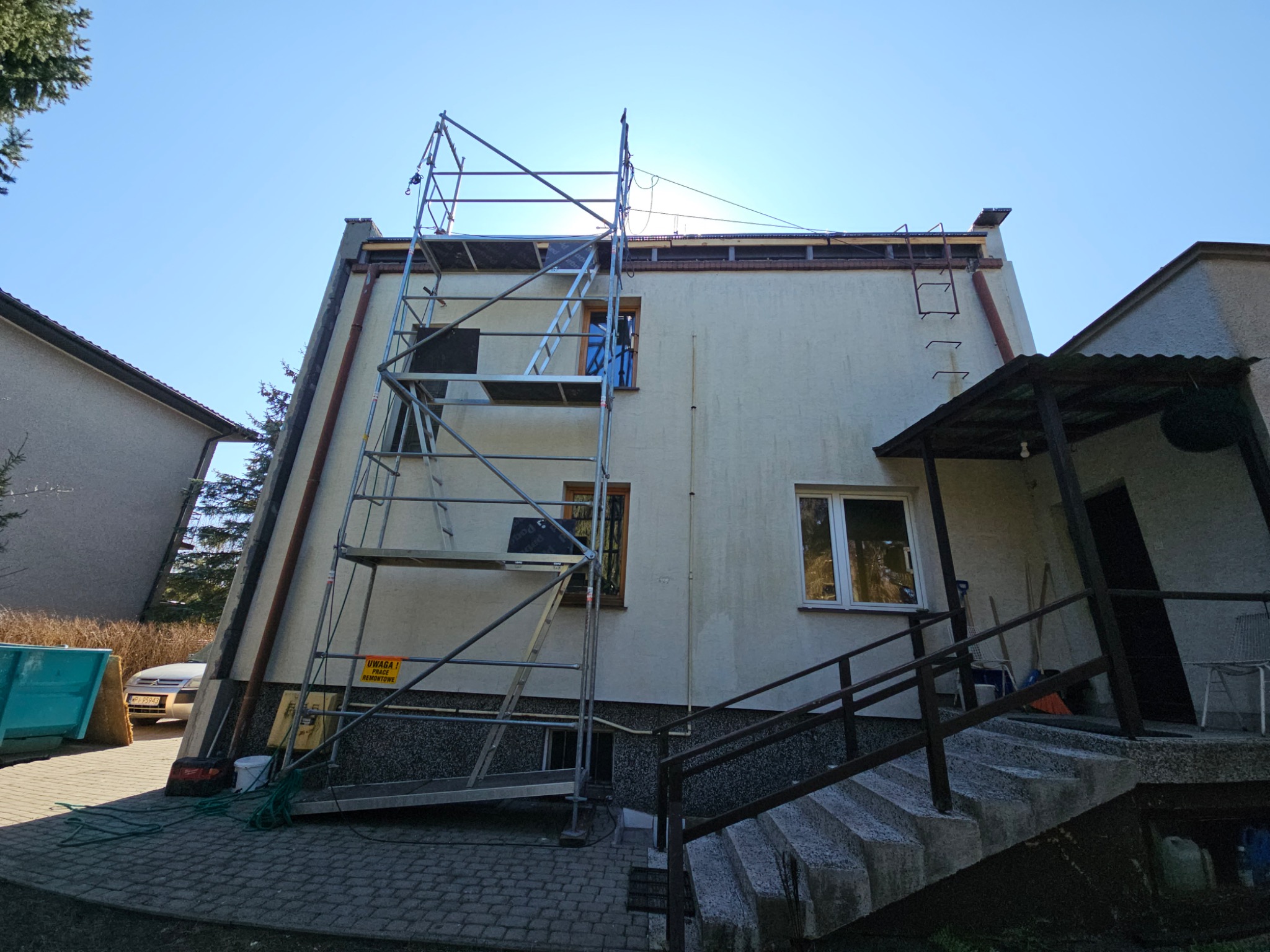 Dom jednorodzinny w trakcie prac remontowych. Widoczne rusztowanie, kontener na gruz i fragment elewacji z oknami. Słoneczny dzień.