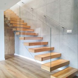 Glasco Sp. z o.o. - Nowoczesne schody z drewnianymi stopniami, szklaną balustradą i metalową poręczą, na tle ściany z szarych płytek. Widoczny fragment podłogi z paneli.