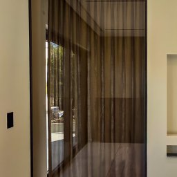 Glasco Sp. z o.o. - Nowoczesne drzwi z ciemnego szkła z czarną ramą, przez które widać zasłony i fragment ogrodu. Minimalistyczny design, jasne ściany i podłoga.