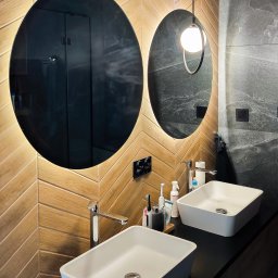 Glasco Sp. z o.o. - Nowoczesna łazienka z dwoma owalnymi lustrami podświetlanymi LED, umywalkami nablatowymi i drewnopodobną okładziną ścienną ułożoną w jodełkę.