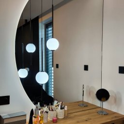 Glasco Sp. z o.o. - Owalne lustro z podświetleniem LED nad drewnianą toaletką, odbijające wiszące lampy kuliste. Widoczne kosmetyki i akcesoria do makijażu. Minimalistyczny design.