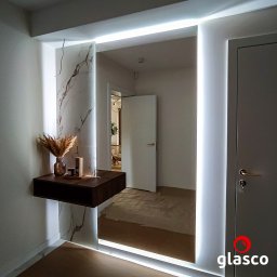 Glasco Sp. z o.o. - Nowoczesne lustro z podświetleniem LED w przedpokoju, obok drzwi i wiszącej półki z dekoracjami. Marmurowy wzór na ścianie dodaje elegancji.