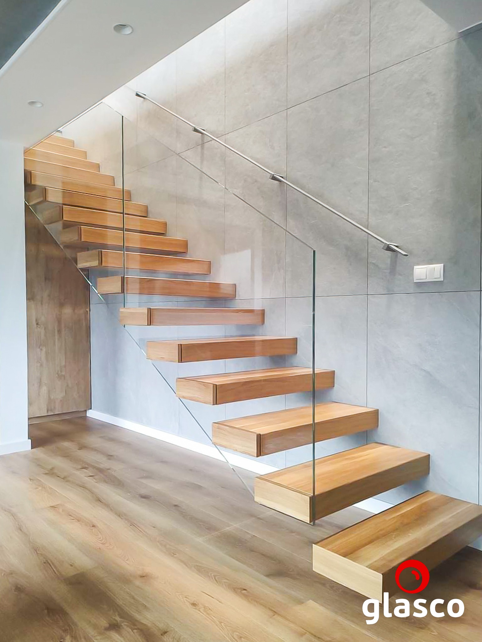 Drewniane schody wspornikowe z przezroczystą taflą szkła jako balustradą i metalową poręczą, zamocowaną do ściany z szarych, kwadratowych płytek.