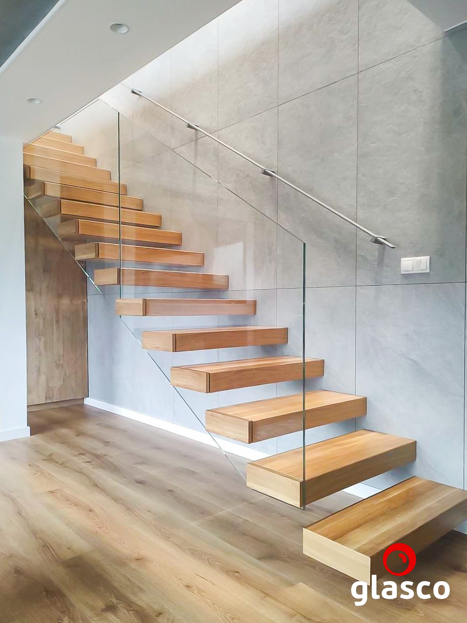 Nowoczesne schody z drewnianymi stopniami, szklaną balustradą i metalową poręczą, na tle ściany z szarych płytek. Widoczny fragment podłogi z paneli.