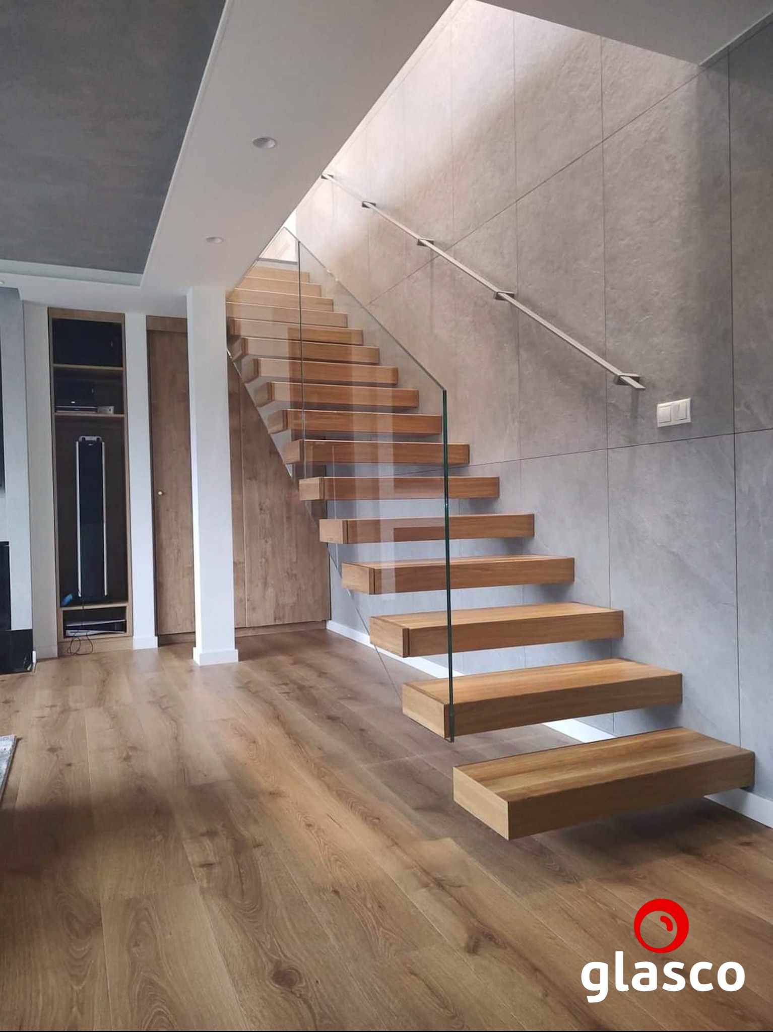 Nowoczesne schody z drewnianymi stopniami, szklaną balustradą i metalową poręczą, w jasnym wnętrzu z drewnianą podłogą i szarą ścianą. Minimalistyczny design.