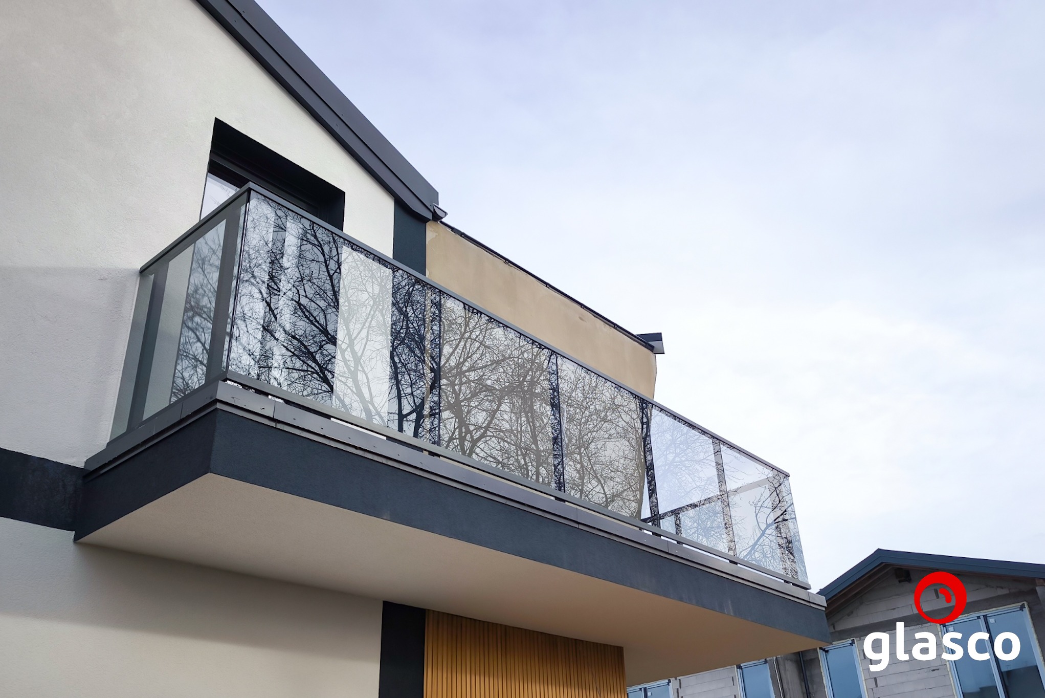 Nowoczesny balkon z przeszkloną balustradą, odbijającą korony drzew. Minimalistyczny design, szara kolorystyka, widok z dołu na tle jasnego nieba.