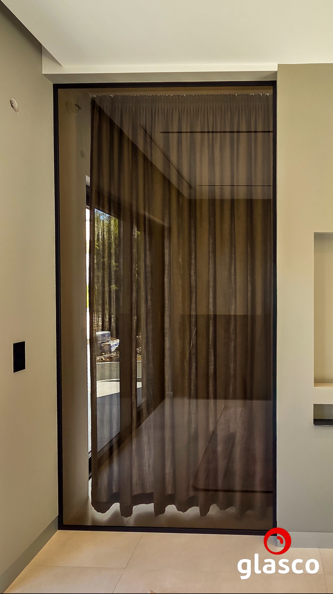 Nowoczesne drzwi z ciemnego szkła z czarną ramą, przez które widać zasłony i fragment ogrodu. Minimalistyczny design, jasne ściany i podłoga.