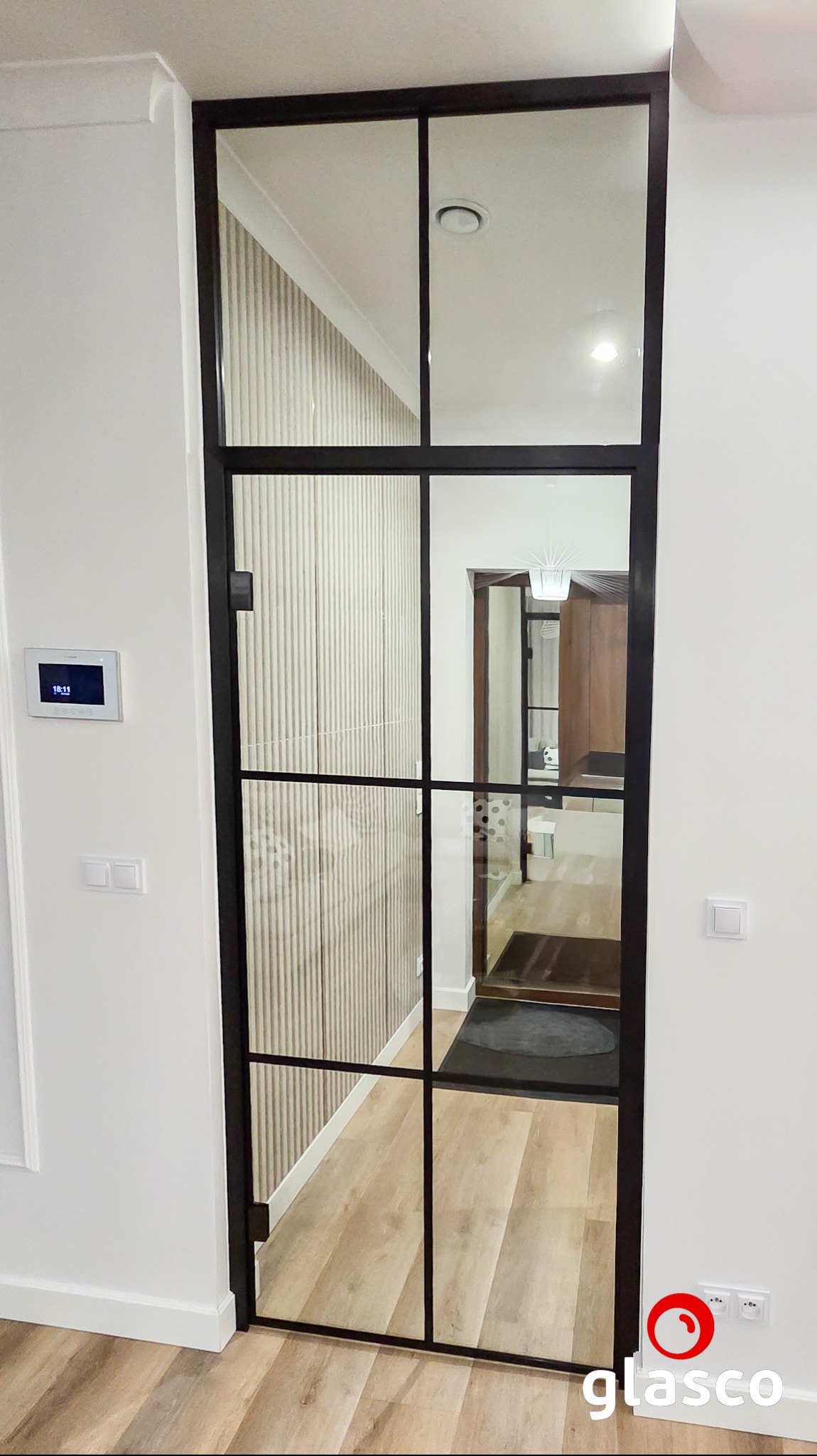 Szklane drzwi w czarnej ramie, z podziałami na prostokąty, oddzielają pomieszczenia. Widoczny fragment korytarza z panelami ściennymi i podłogą.