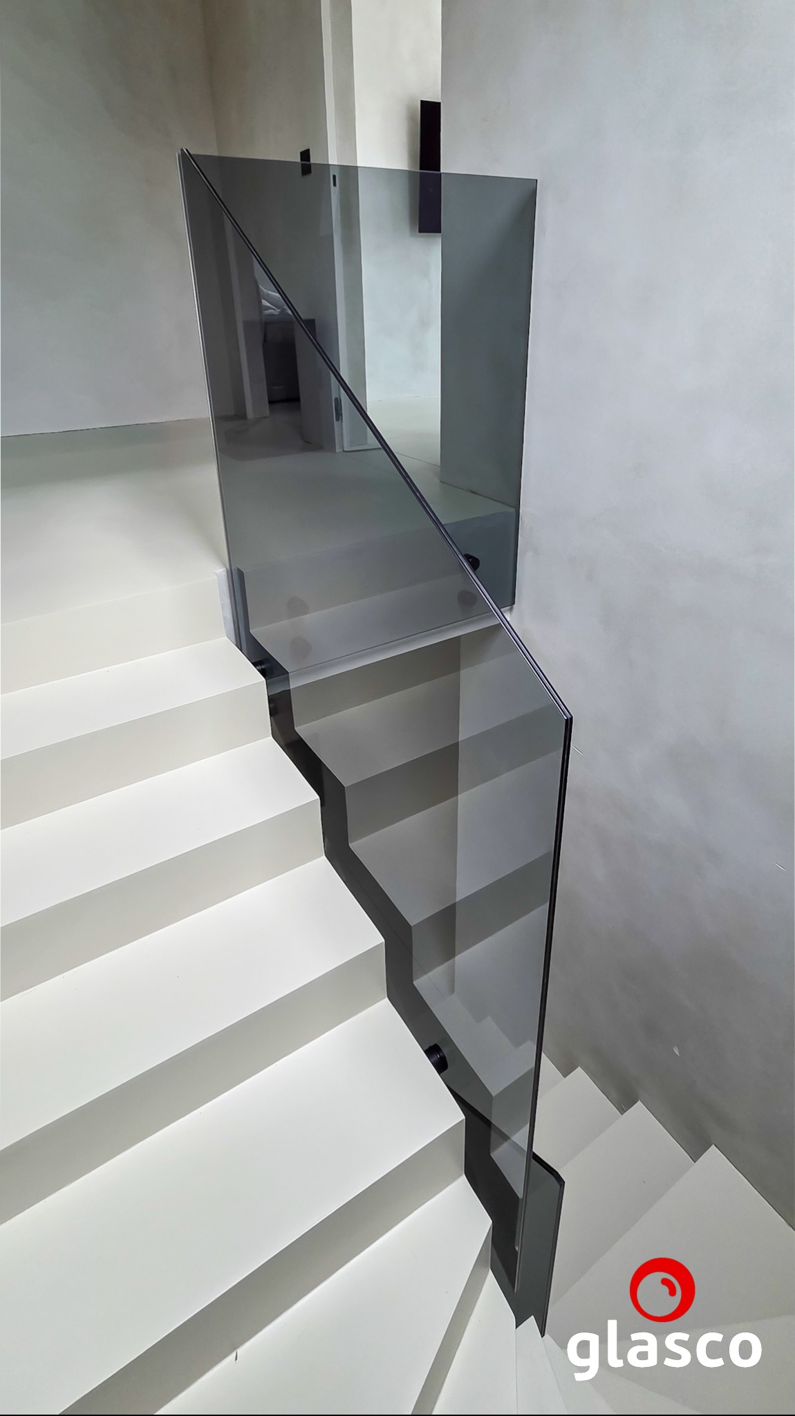 Minimalistyczne schody z szarą, szklaną balustradą. Nowoczesny design, neutralna kolorystyka ścian. Widok z góry na dół, logo firmy w prawym dolnym rogu.