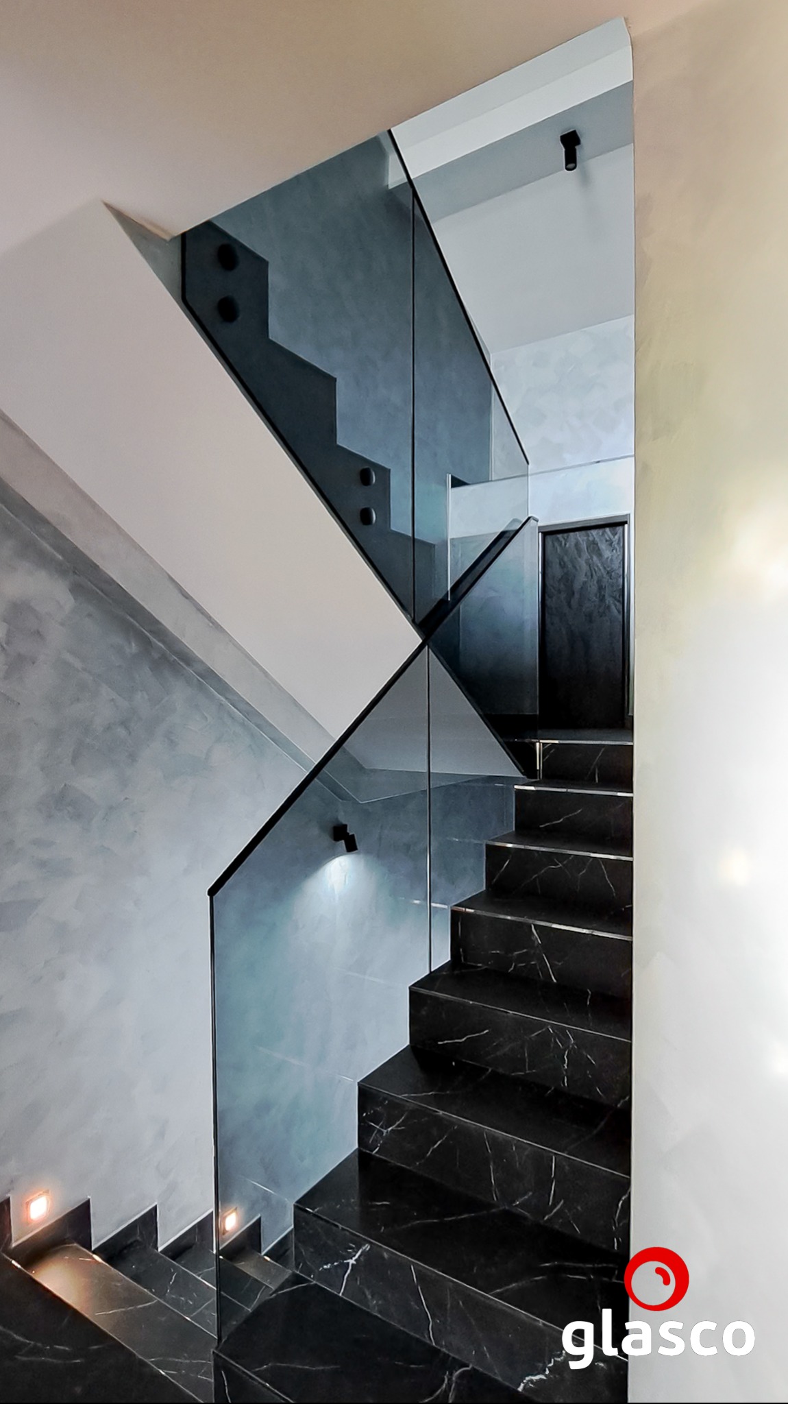 Nowoczesne schody z czarnego marmuru z balustradą ze szkła hartowanego, mocowaną punktowo. Minimalistyczne oświetlenie schodowe i sufitowe. Eleganckie wykończenie wnętrza.