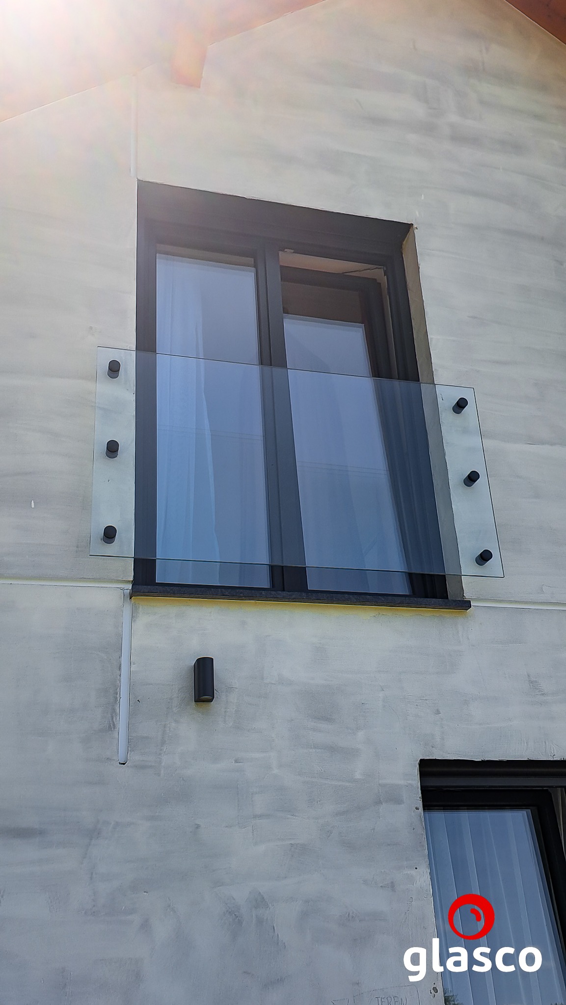 Szklana balustrada balkonowa, mocowana punktowo do ściany budynku o surowym wykończeniu. Widoczne okno z czarną ramą i lampa ścienna. Słoneczny dzień.