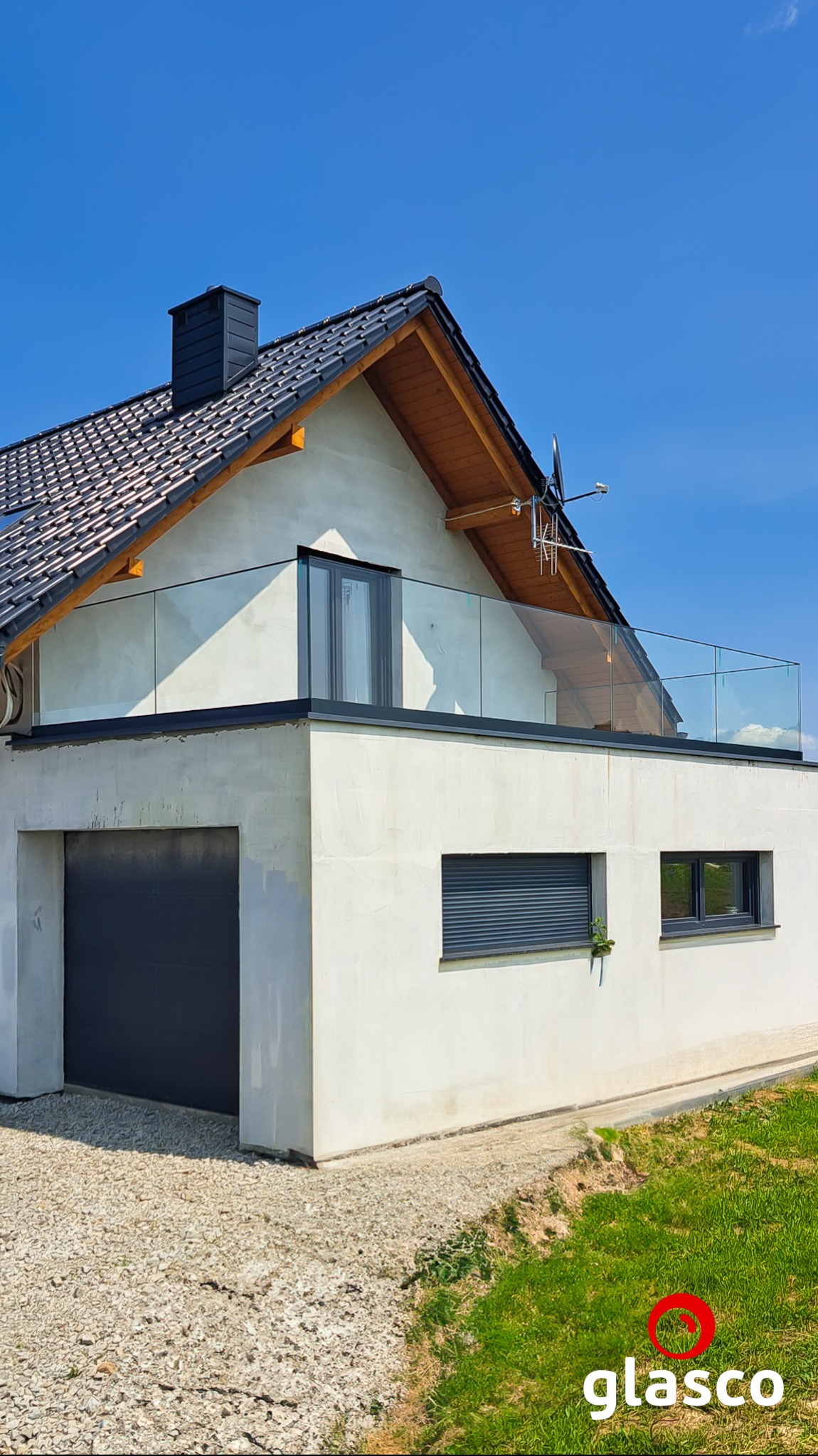 Nowoczesny dom z balkonem zabezpieczonym szkłem, widok z dołu. Minimalistyczny design, jasna elewacja, czarne okna i dachówka. Słoneczny dzień.