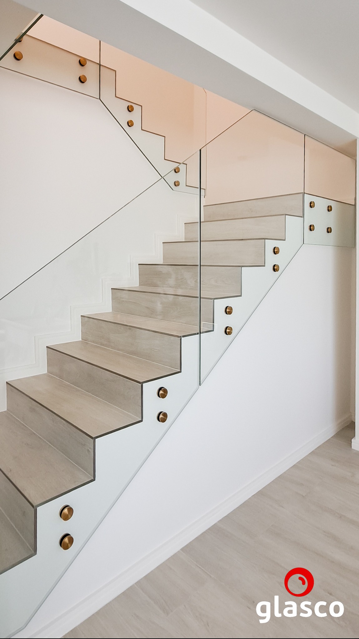 Nowoczesne schody z drewnianymi stopniami i szklaną balustradą mocowaną na złotych uchwytach. Minimalistyczny design, jasne wnętrze, widok z dołu na górę.