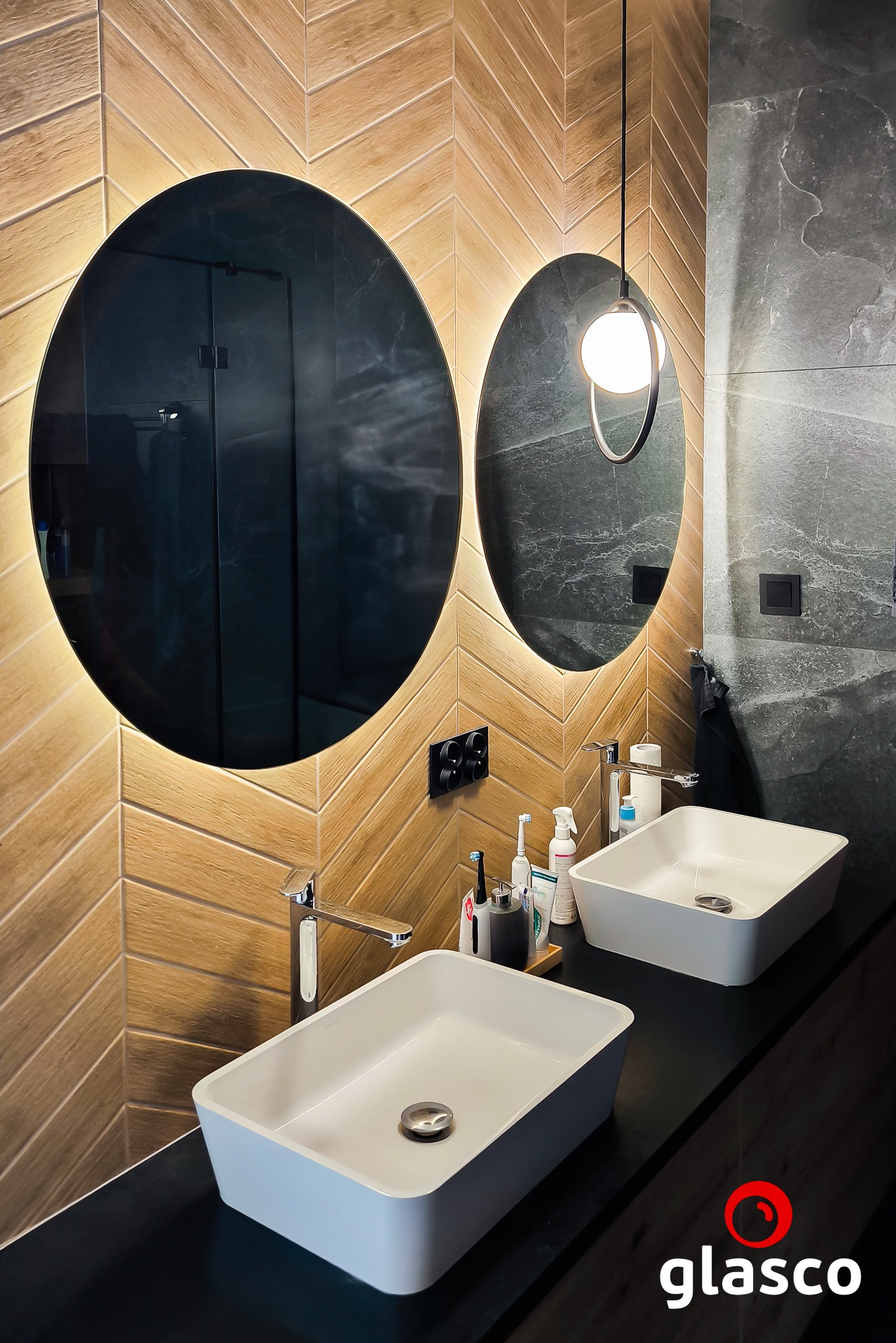 Nowoczesna łazienka z dwoma owalnymi lustrami podświetlanymi LED, umywalkami nablatowymi i drewnopodobną okładziną ścienną ułożoną w jodełkę.