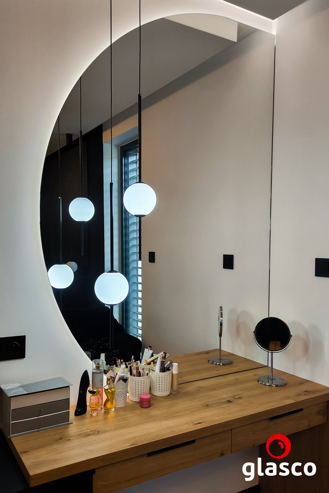 Owalne lustro z podświetleniem LED nad drewnianą toaletką, odbijające wiszące lampy kuliste. Widoczne kosmetyki i akcesoria do makijażu. Minimalistyczny design.