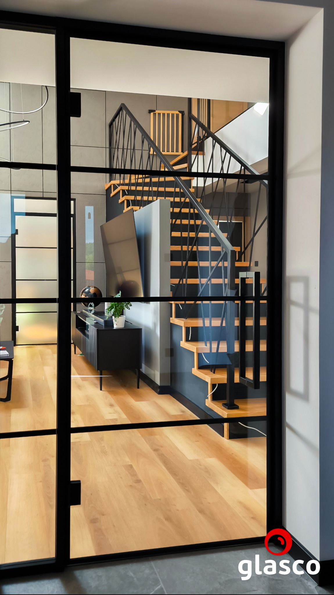 Szklane drzwi z czarnymi szprosami, widok na schody z metalową balustradą i drewnianymi stopniami. W tle salon z szafką RTV i telewizorem.