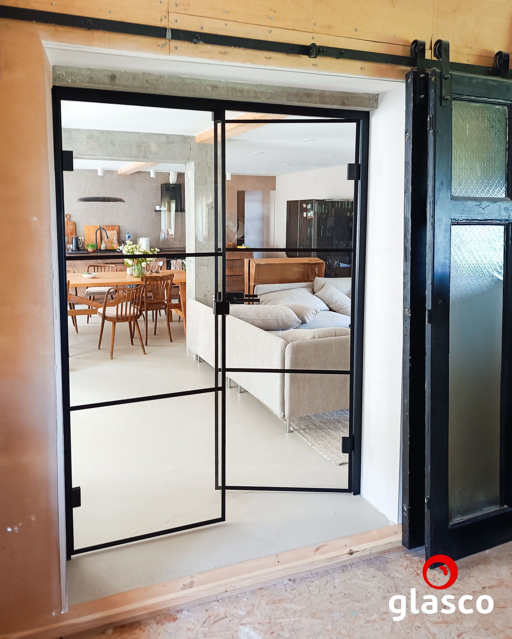 Nowoczesne, dwuskrzydłowe drzwi szklane z czarnymi szprosami, oddzielające kuchnię od salonu. Widoczny system przesuwny i fragment starych drzwi z szybą ornamentową.