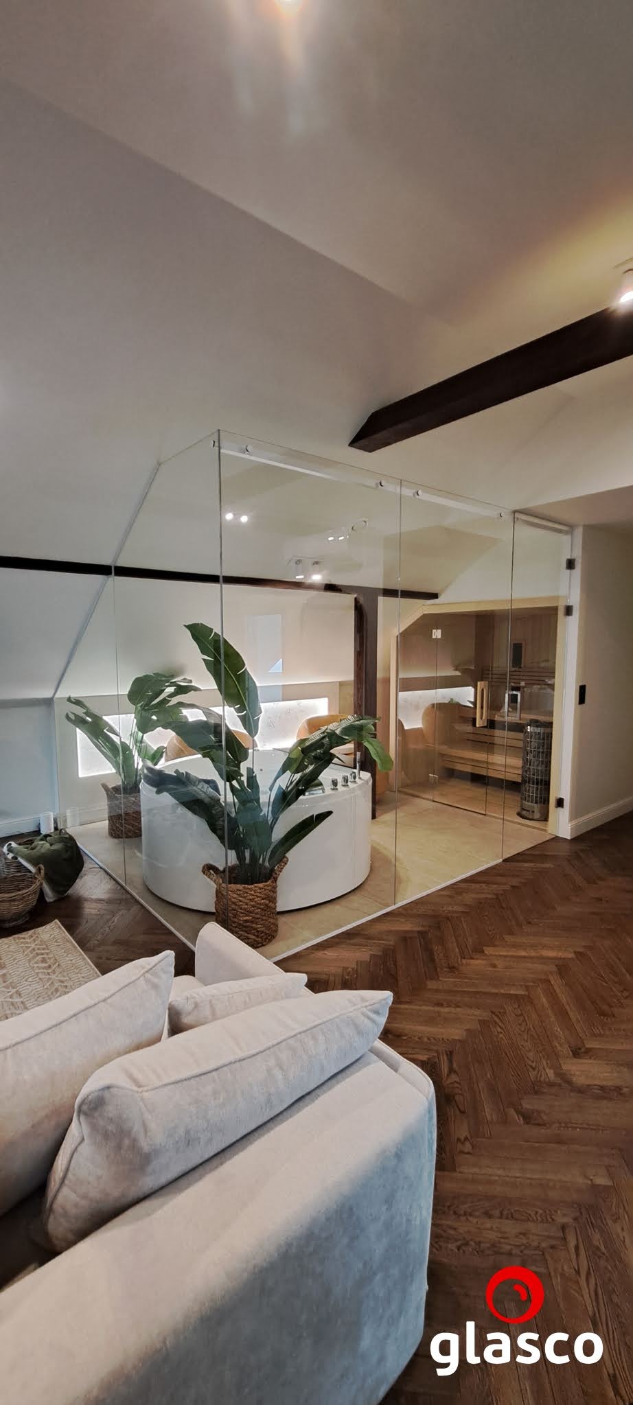 Szklana zabudowa w łazience z sauną, widok z salonu. Przezroczyste ściany oddzielają strefę relaksu. Parkiet w jodełkę na podłodze, rośliny w koszach.