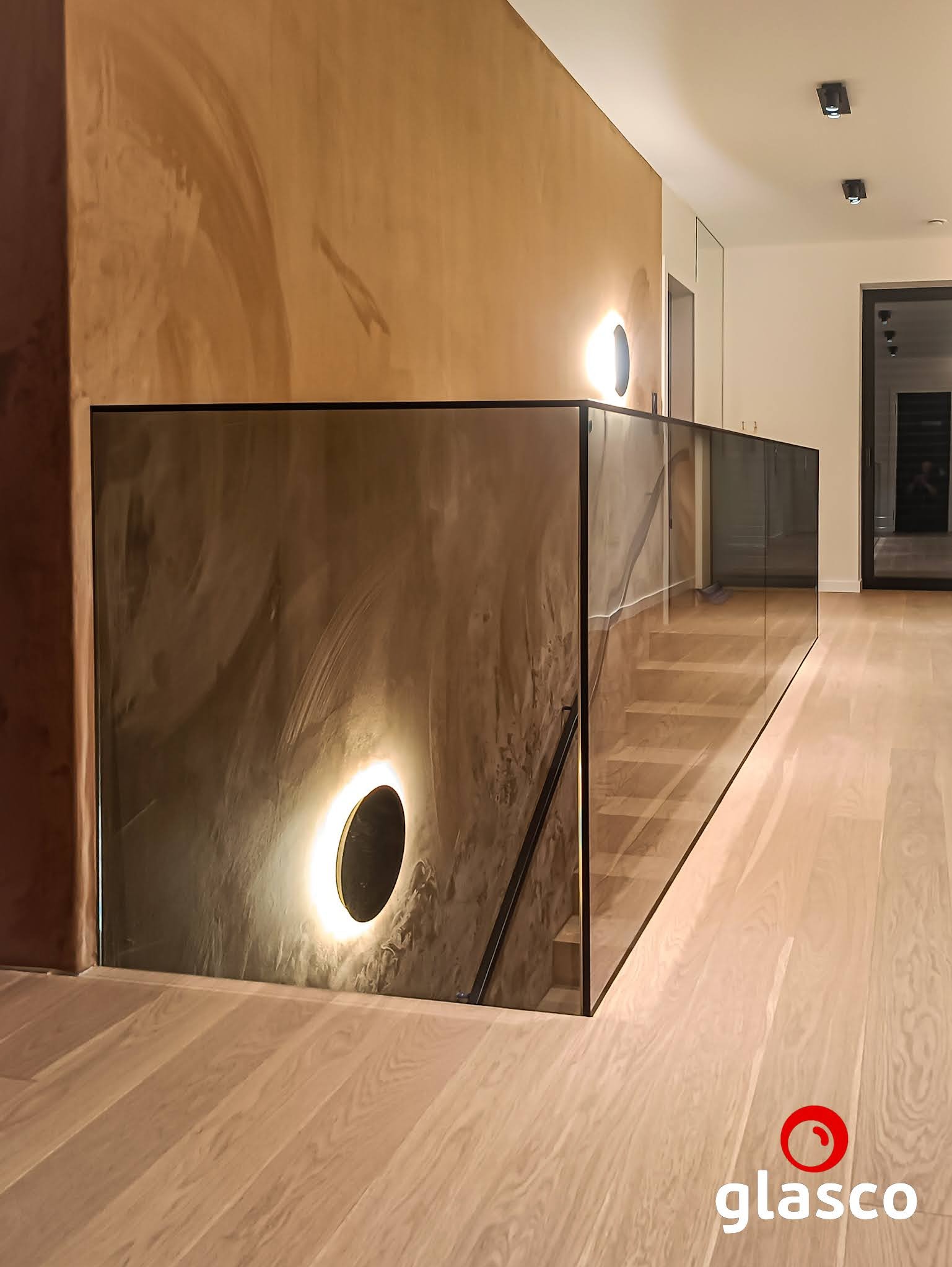 Nowoczesna balustrada ze szkła, z czarną ramą i poręczą, oświetlona okrągłym kinkietem. Widoczne drewniane schody i podłoga. Minimalistyczny design.