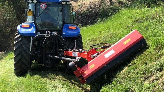 Niebieski traktor z czerwoną kosiarką bijakową na zboczu, świeżo skoszona trawa, widok z tyłu, tablica rejestracyjna BM-2402, znak ograniczenia prędkości 40 km/h.