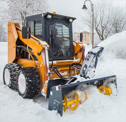 Żółta frezarka do śniegu w akcji, wyrzucająca śnieg na bok. Widoczne drzewa i budynek w tle, pokryte śniegiem. Zimowy krajobraz.
