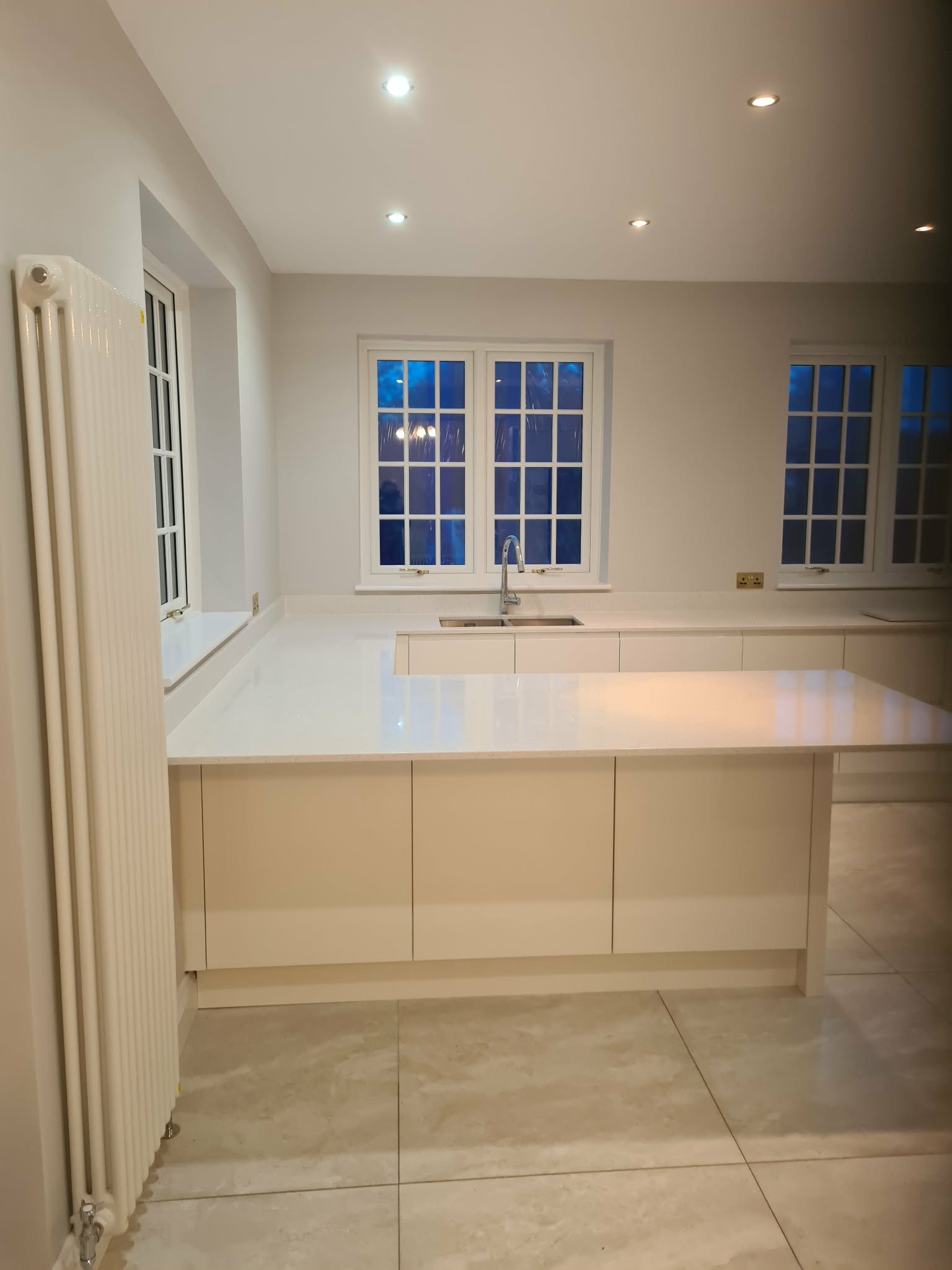 Jasna kuchnia z wyspą i oknami z widokiem na nocne niebo. Minimalistyczny design, kremowe szafki, biały blat i płytki podłogowe. Grzejnik dekoracyjny przy ścianie.