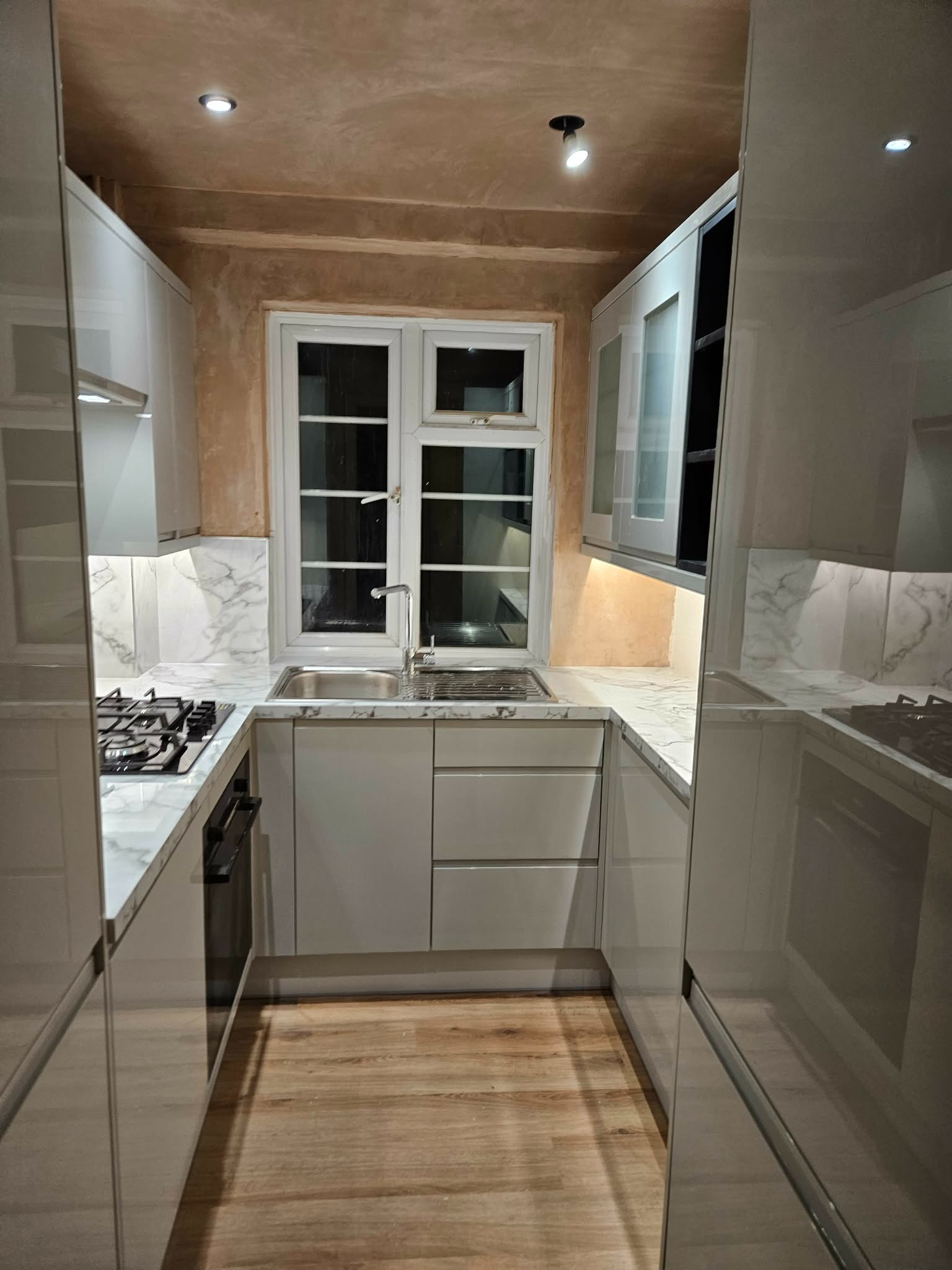 Nowoczesna kuchnia w trakcie wykończenia: białe, błyszczące szafki, marmurowy blat, drewniana podłoga, surowy tynk na suficie i ścianie nad oknem. Widok na okno.