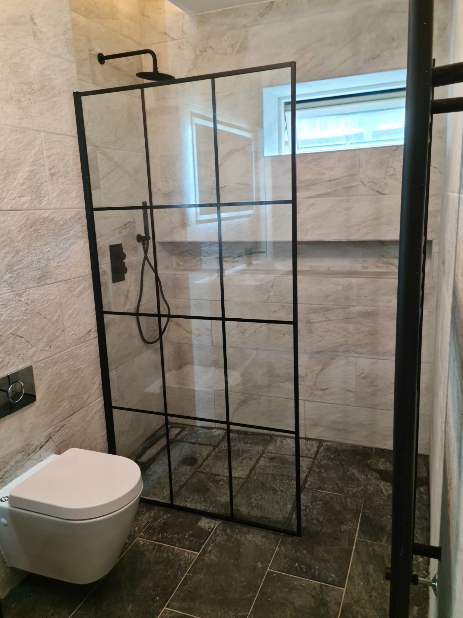 Nowoczesna kabina prysznicowa z czarnymi profilami i szklanymi ściankami, zamontowana w wykończonej łazience z beżowymi płytkami i ciemną podłogą. Widoczna toaleta podwieszana.