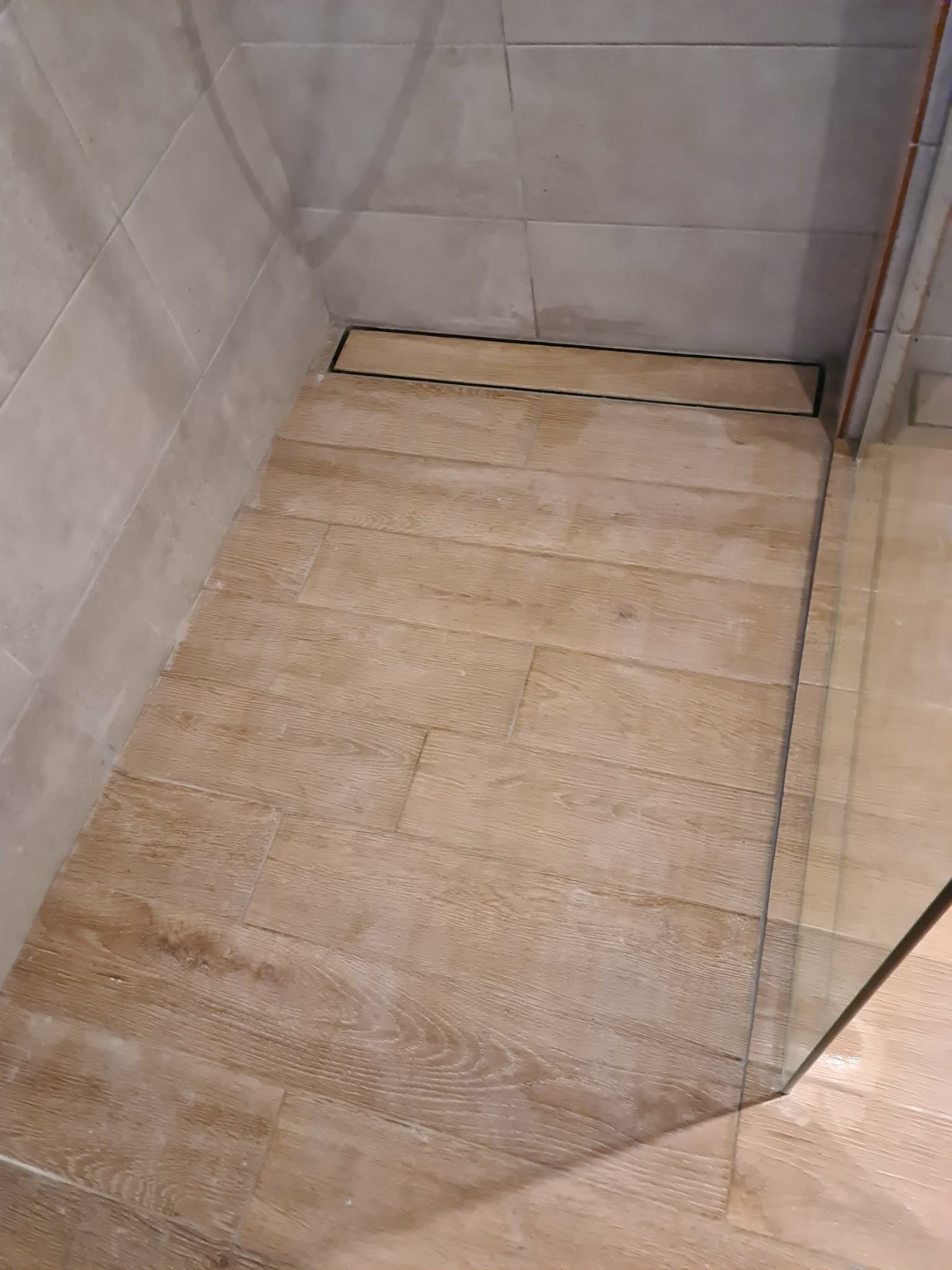 Wykończona strefa prysznicowa z odpływem liniowym i płytkami imitującymi drewno na podłodze, oraz jasnymi płytkami na ścianach. Widoczny fragment szklanej kabiny.