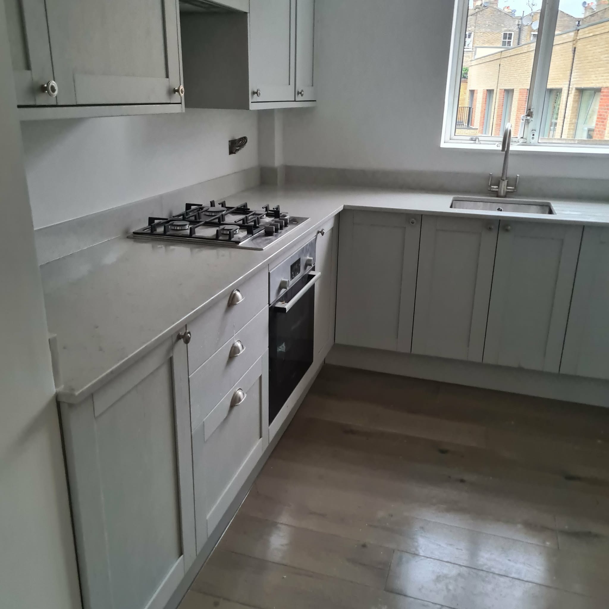 Jasna kuchnia w stylu angielskim z drewnianą podłogą, widok na okno z zabudową miejską. Uwagę przykuwają frezowane fronty szafek i kamienny blat.