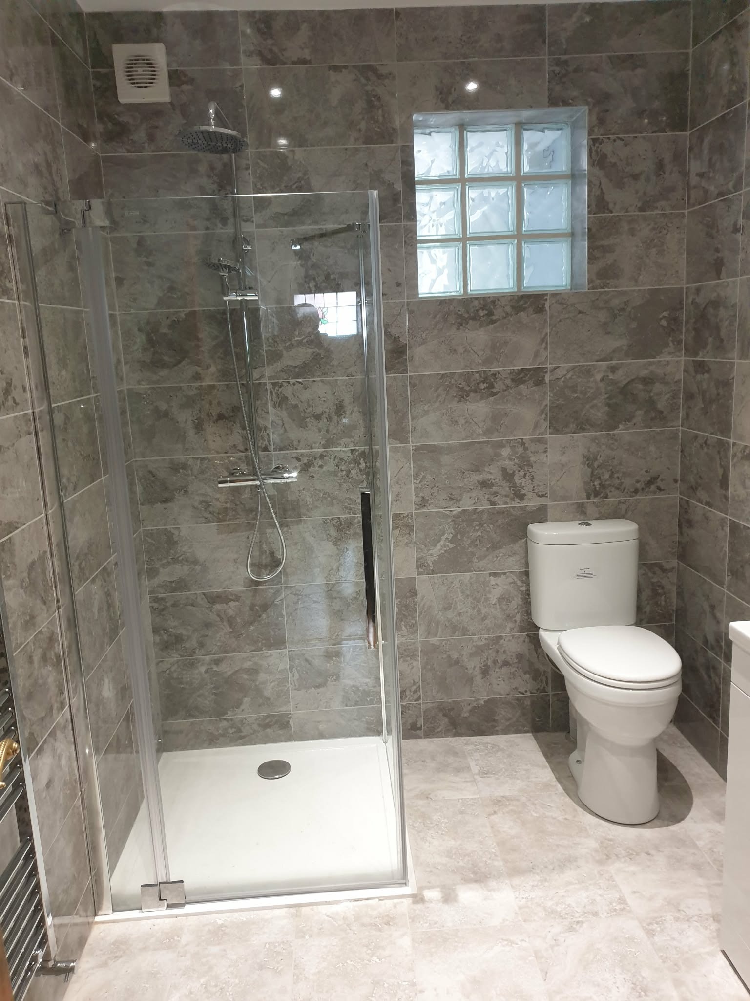 Nowoczesna łazienka z kabiną prysznicową, toaletą i szarymi płytkami imitującymi marmur. Okno z luksferów wpuszcza naturalne światło. Minimalistyczny design.