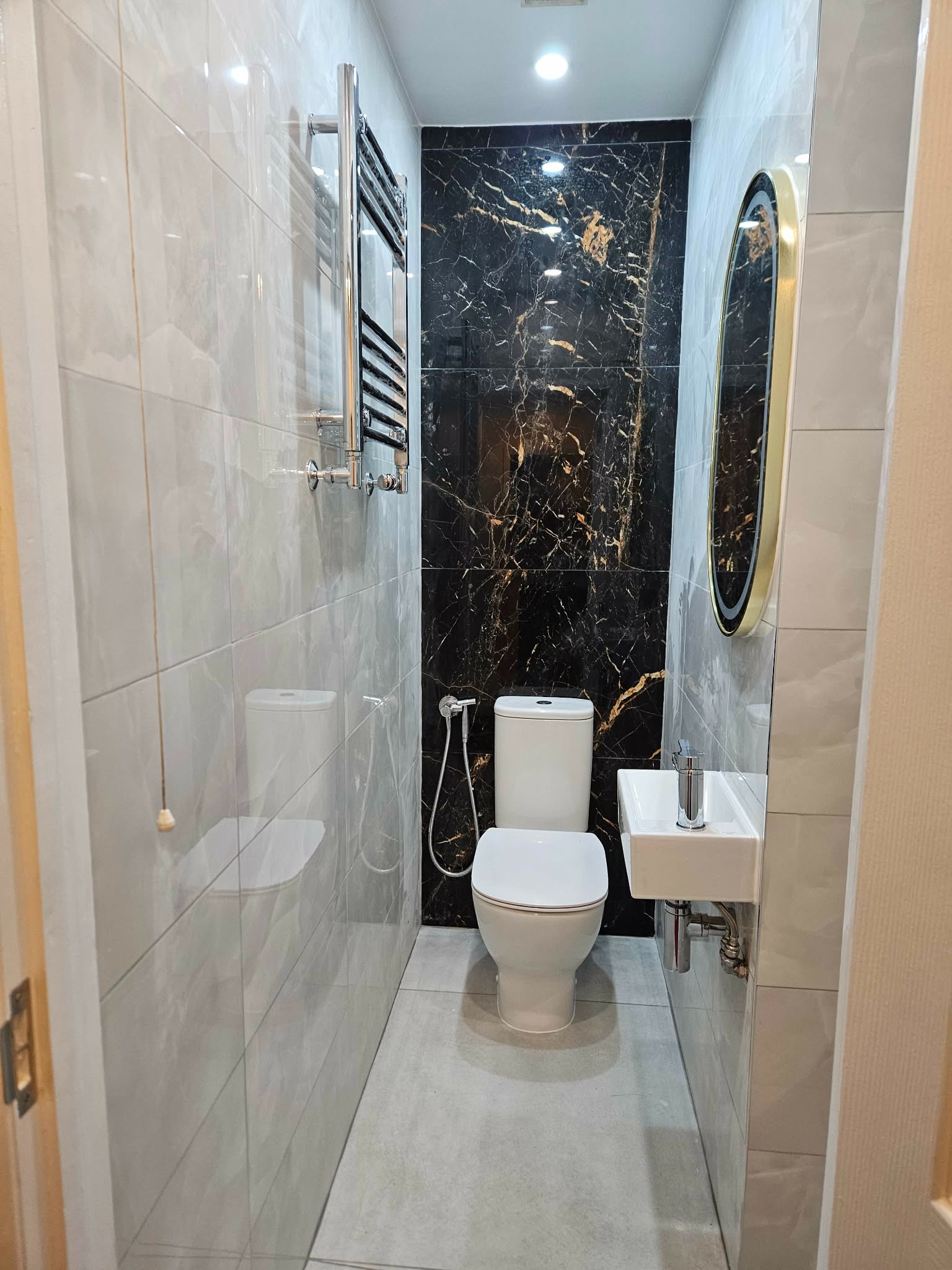 Wykończona łazienka z toaletą, umywalką i grzejnikiem na ścianie z szarych płytek. Jedna ściana wykończona czarnym marmurem. Nad umywalką owalne lustro w złotej ramie.