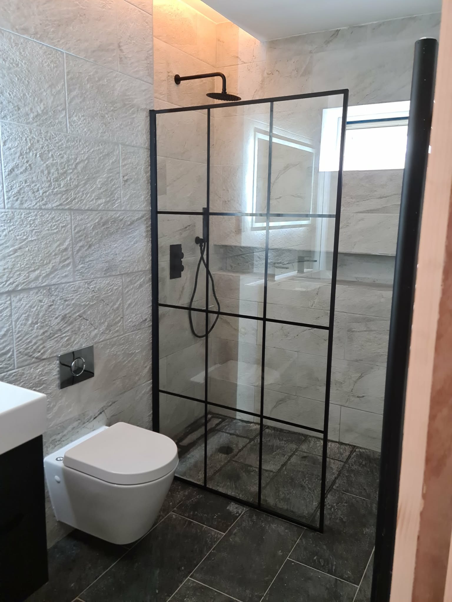 Nowoczesna łazienka po remoncie: kabina prysznicowa z czarnymi profilami, podwieszana toaleta, płytki imitujące kamień na ścianach i podłodze. Minimalistyczny design.