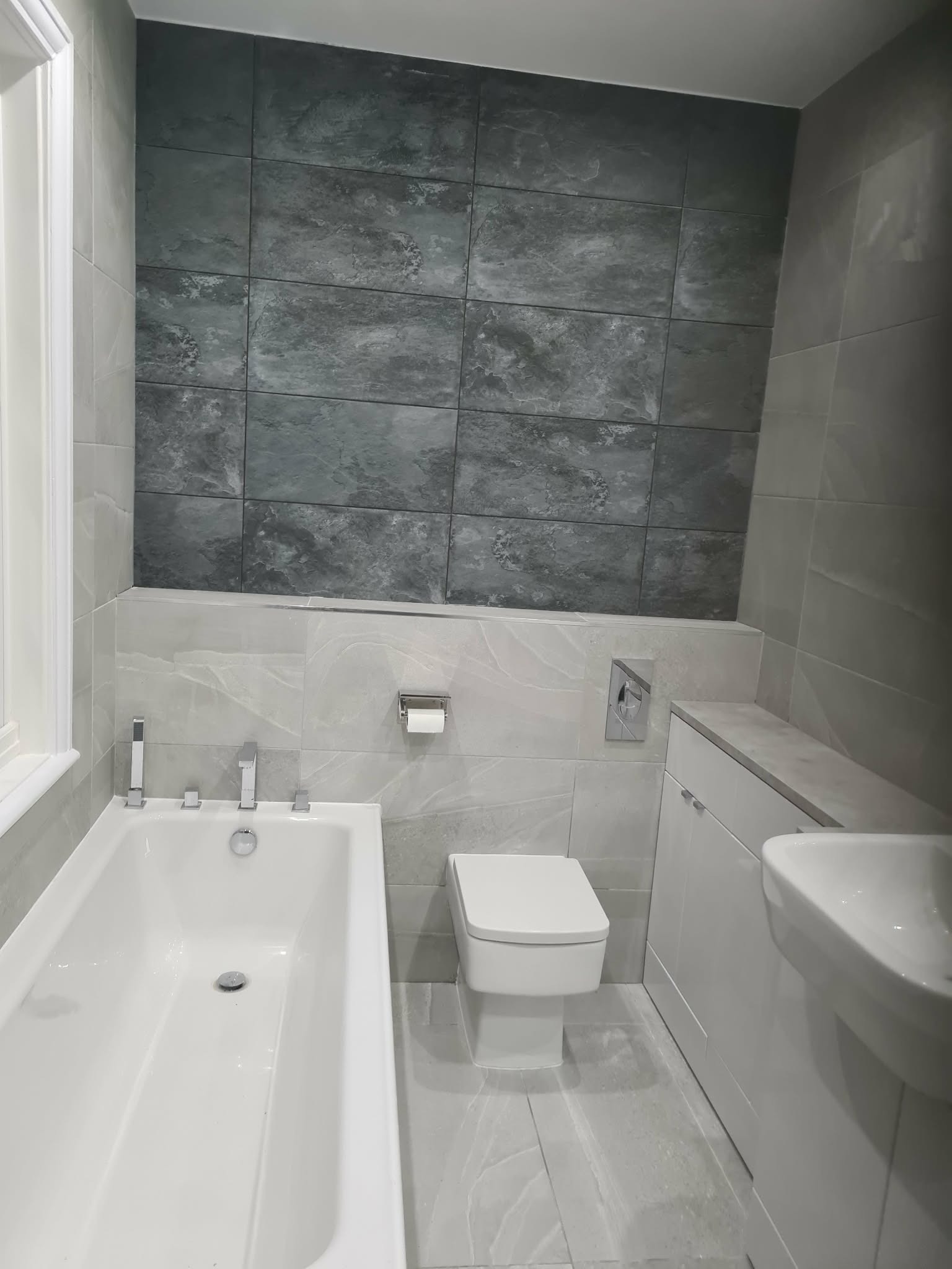 Wykończona łazienka w Gliwicach: biała wanna, toaleta i umywalka z szafką. Ściany wyłożone szarymi płytkami o zróżnicowanej fakturze, tworząc nowoczesny design.