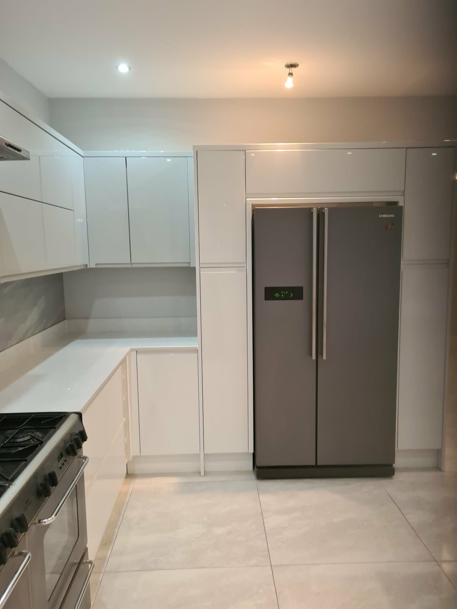 Nowoczesna, jasna kuchnia z białymi, lakierowanymi frontami szafek, szarym blatem i dużą, dwudrzwiową lodówką Samsung w zabudowie. Minimalistyczny design.