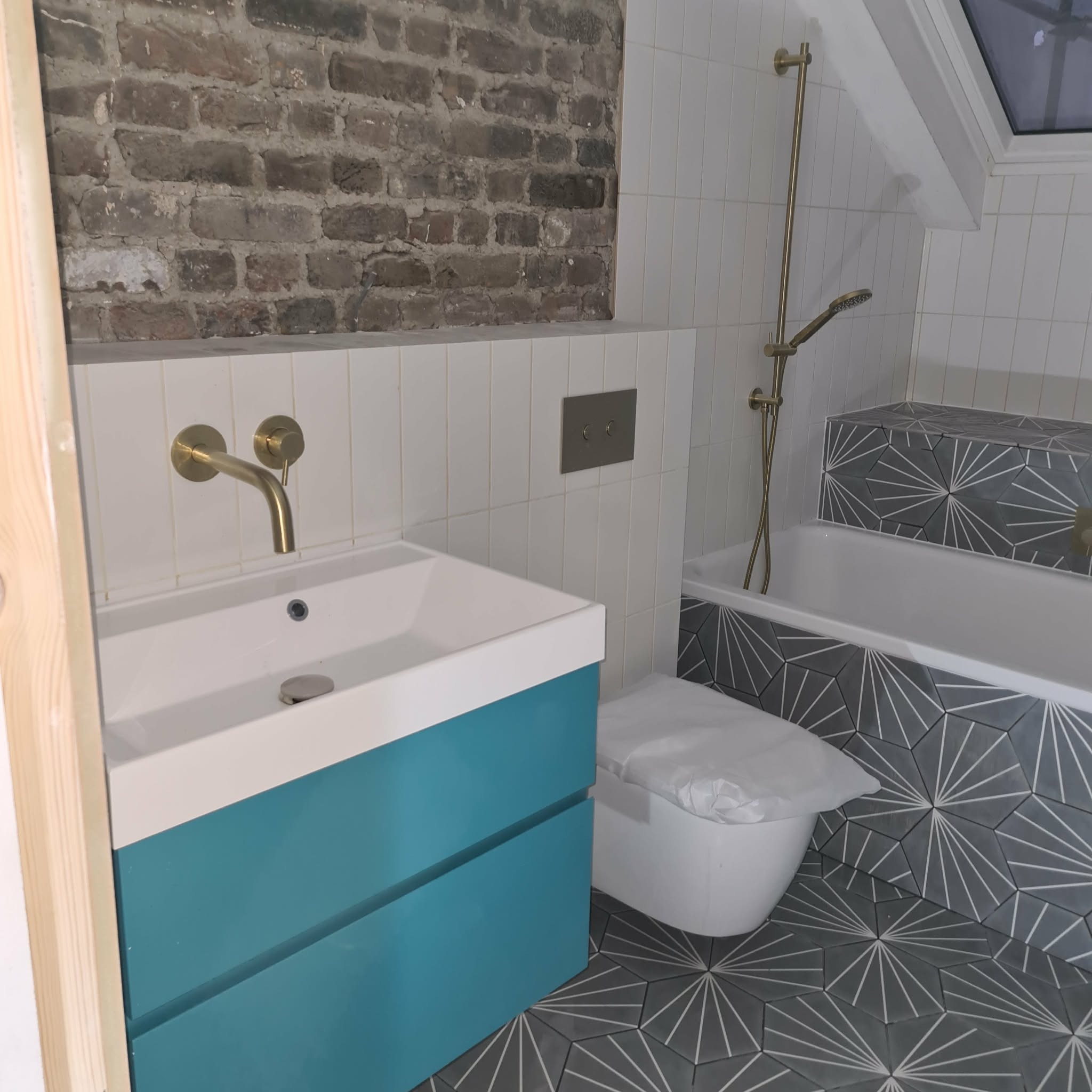 Nowoczesna łazienka z turkusową szafką pod umywalką, złotą armaturą i szarą mozaiką na podłodze i obudowie wanny. Częściowo widoczna ściana z cegły.