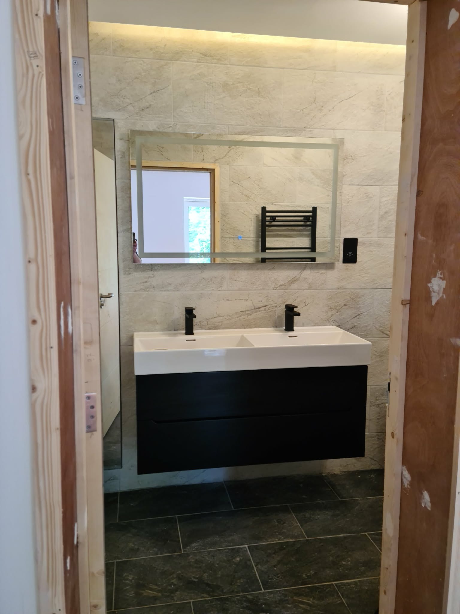 Nowoczesna łazienka z podwójną umywalką nablatową, czarną szafką pod umywalką i podświetlanym lustrem. Widok przez surową ościeżnicę drzwi.