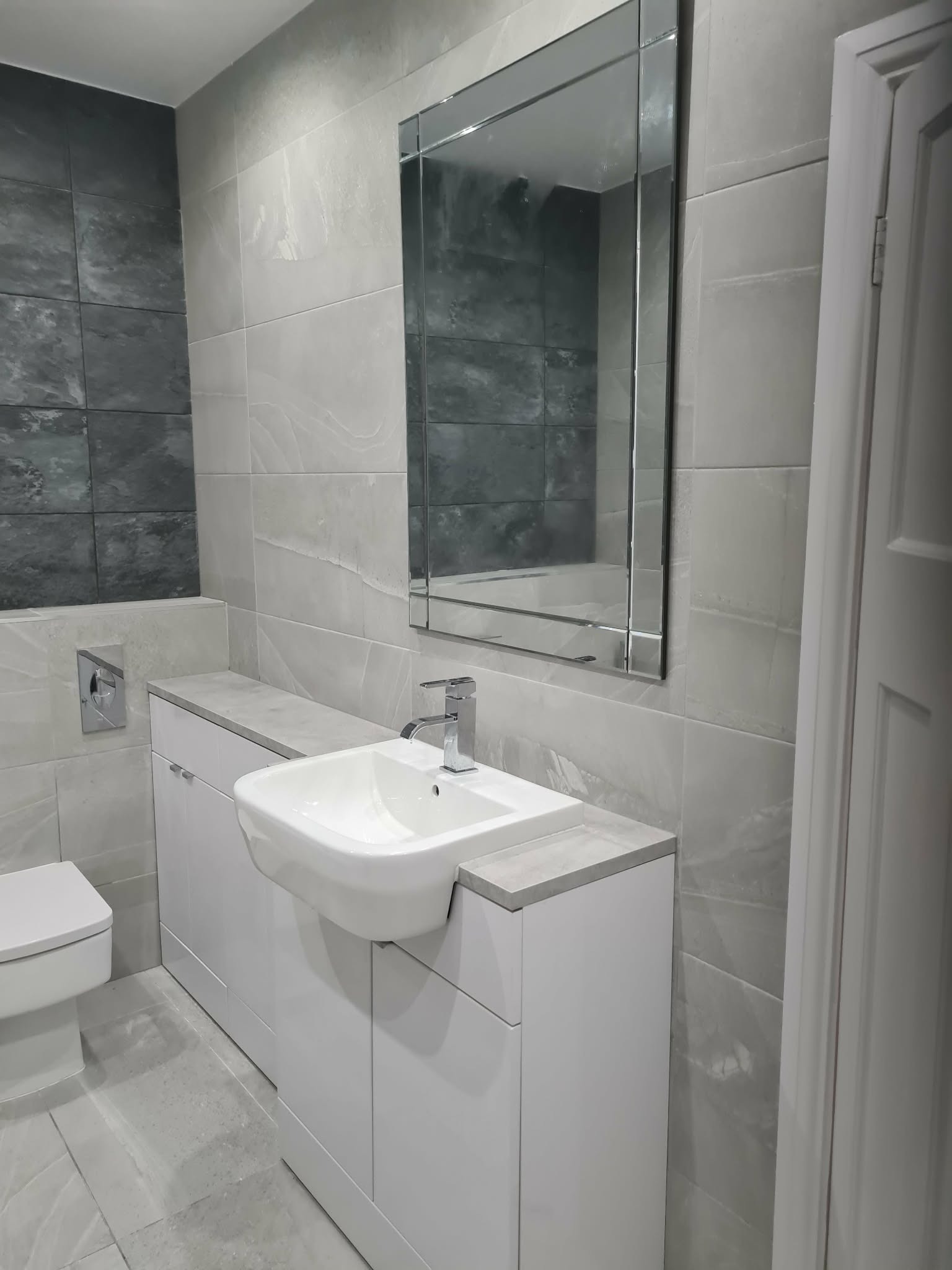 Nowoczesna łazienka w szarościach: biała umywalka z szafką, lustro w ramie, płytki imitujące kamień na ścianach i podłodze. Minimalistyczny design, czyste linie.