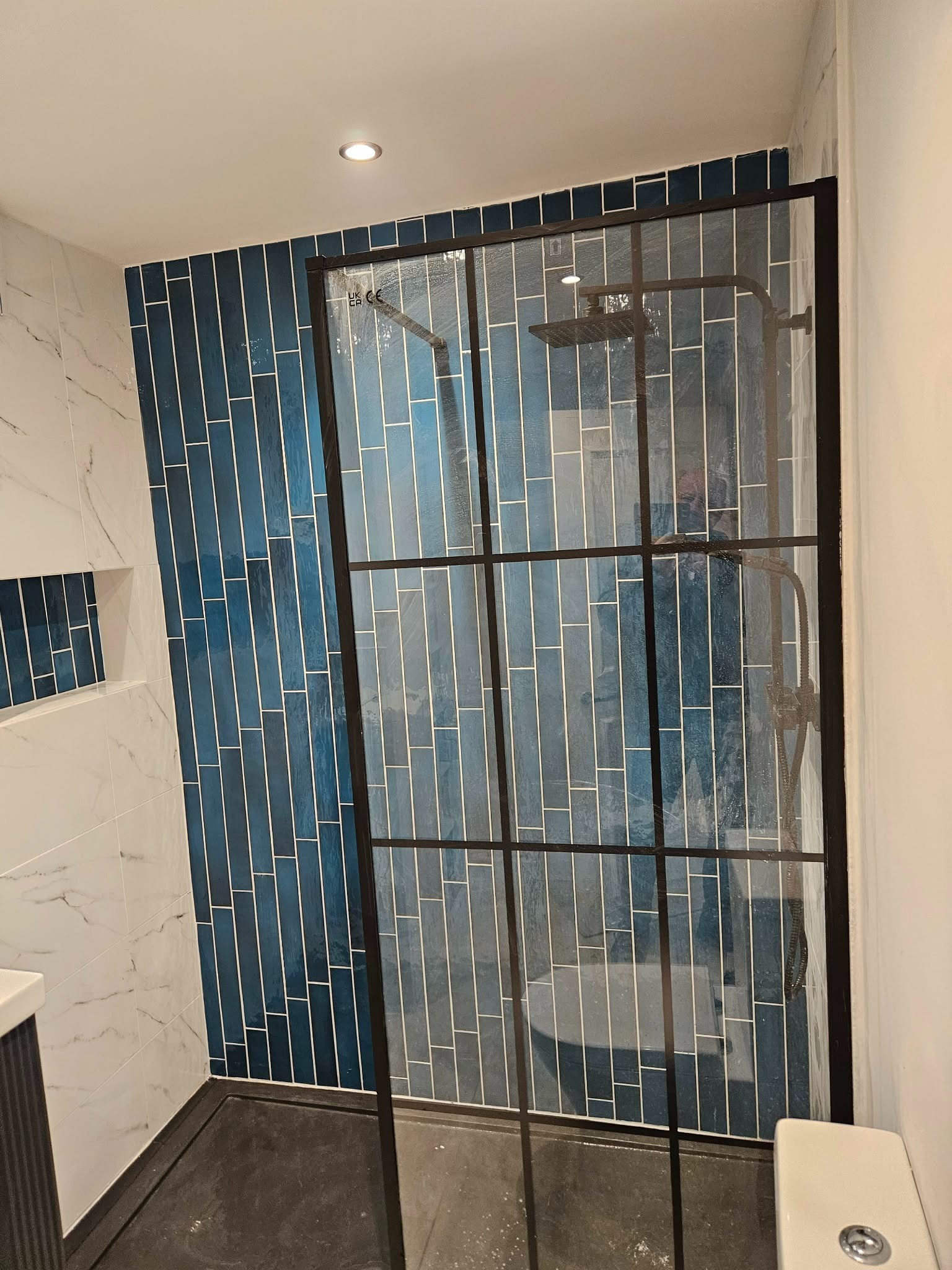 Wykończona łazienka z kabiną prysznicową typu walk-in, wyłożona niebieskimi, pionowymi płytkami i białymi płytkami imitującymi marmur. Czarna rama kabiny.