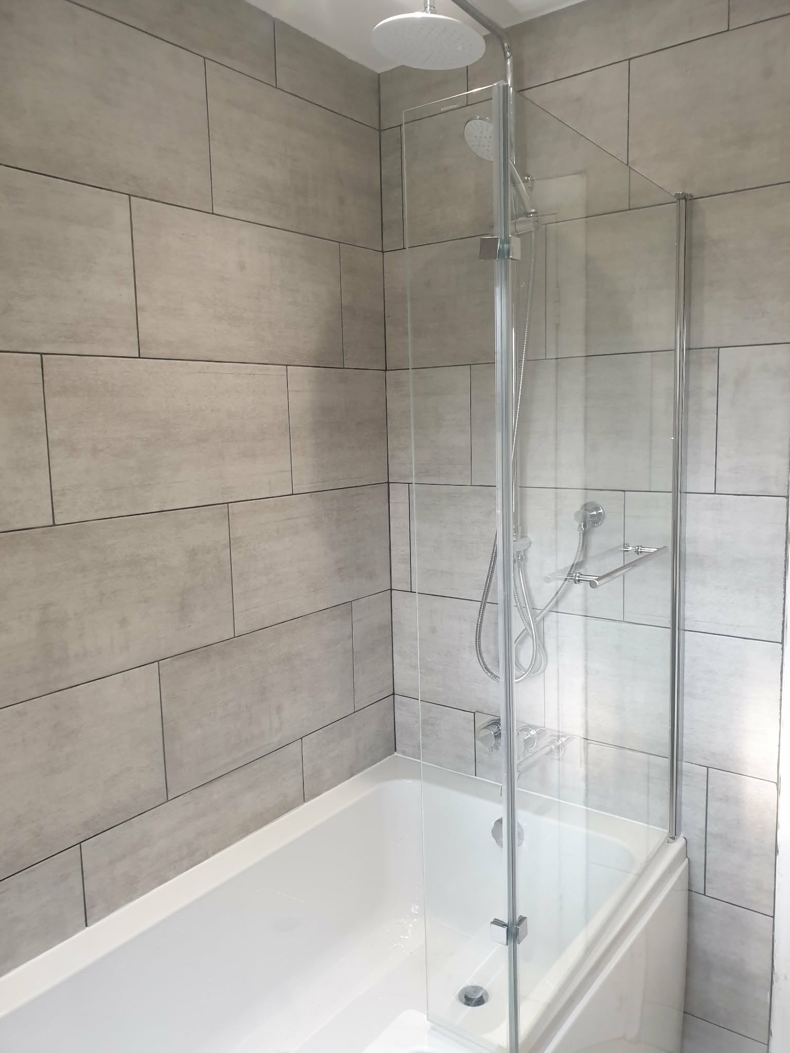 Wykończona łazienka z wanną i szklanym parawanem nawannowym. Ściany wyłożone szarymi, prostokątnymi płytkami. Widoczna deszczownica i bateria prysznicowa.