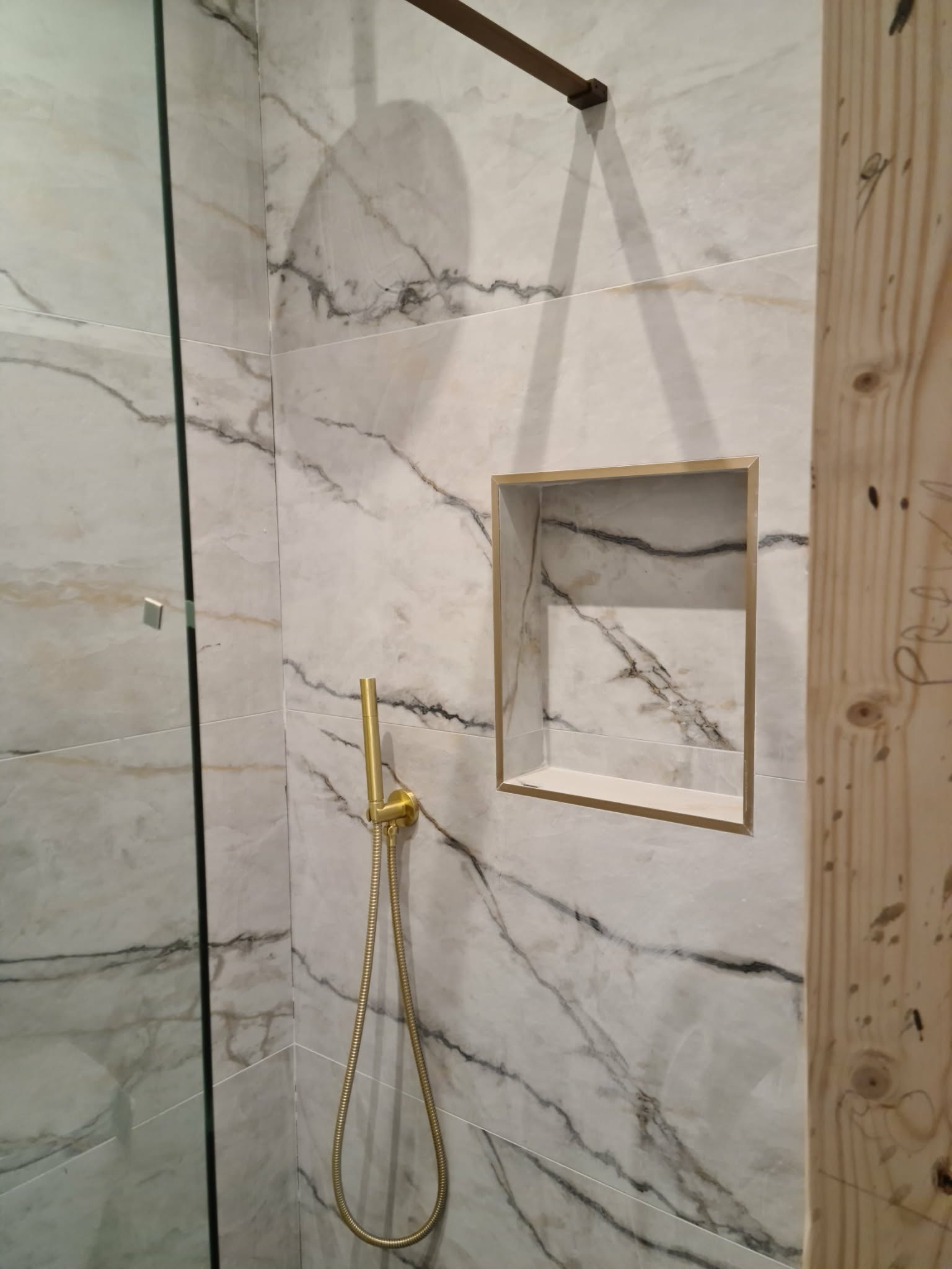 Wykończona kabina prysznicowa z marmurowymi płytkami, złotą armaturą i wnęką. Widoczny fragment drewnianej konstrukcji. Nowoczesny design.