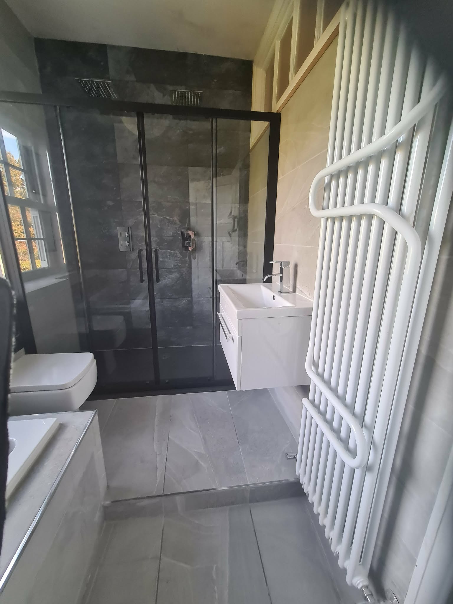 Nowoczesna łazienka z kabiną prysznicową typu walk-in, białą umywalką z szafką i grzejnikiem drabinkowym. Szare płytki podłogowe i ścienne. Czarna rama kabiny.