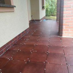 PGbud - Układanie brązowych płytek na tarasie z użyciem krzyżyków dystansowych. Widoczne fragmenty elewacji i ceglanej kolumny. Prace glazurnicze w toku.