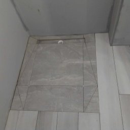 PGbud - Wykończona strefa prysznicowa z odpływem liniowym i szarymi płytkami imitującymi kamień, ułożonymi w różnych formatach. Widok z góry, surowy stan wykończenia.