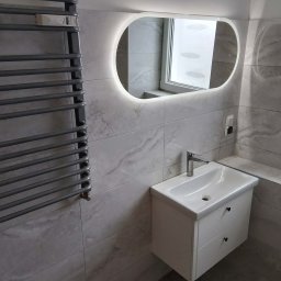 PGbud - Nowoczesna łazienka z szarymi płytkami imitującymi marmur, podświetlanym lustrem owalnym, grzejnikiem drabinkowym i białą szafką z umywalką.